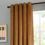 Caramel gold velvet eyelet curtains with brown metal grommets on a dark rod, shown against a light grey wall beside a bright window.