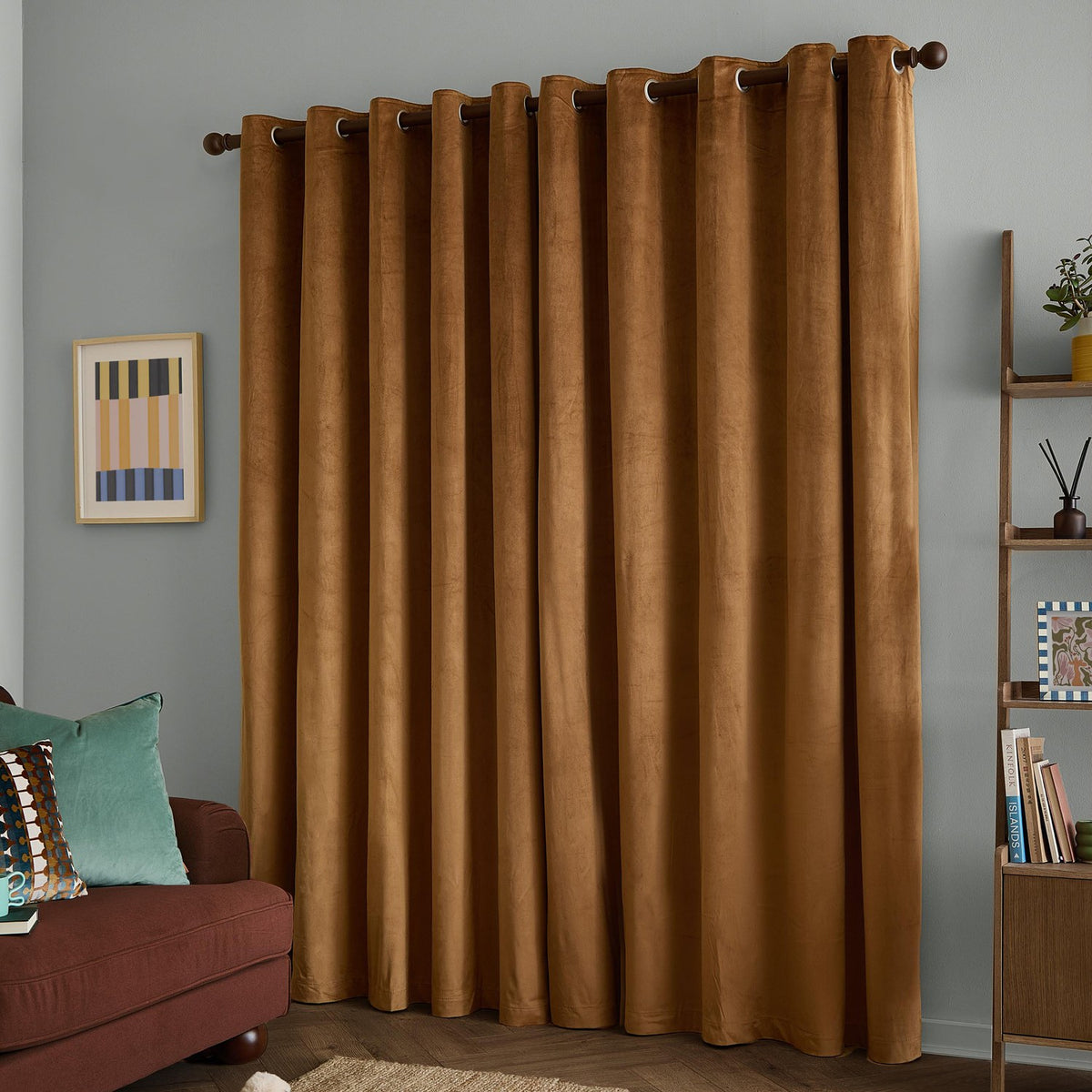 Caramel gold velvet eyelet curtains hanging on a dark wooden pole in a living room, shown with grey walls, framed wall art, and a wooden bookshelf.