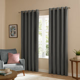 Charcoal grey velvet eyelet curtains with silver grommets hang on a metal rod either side of a window in a living room, styled with a blue armchair and pink cushion.