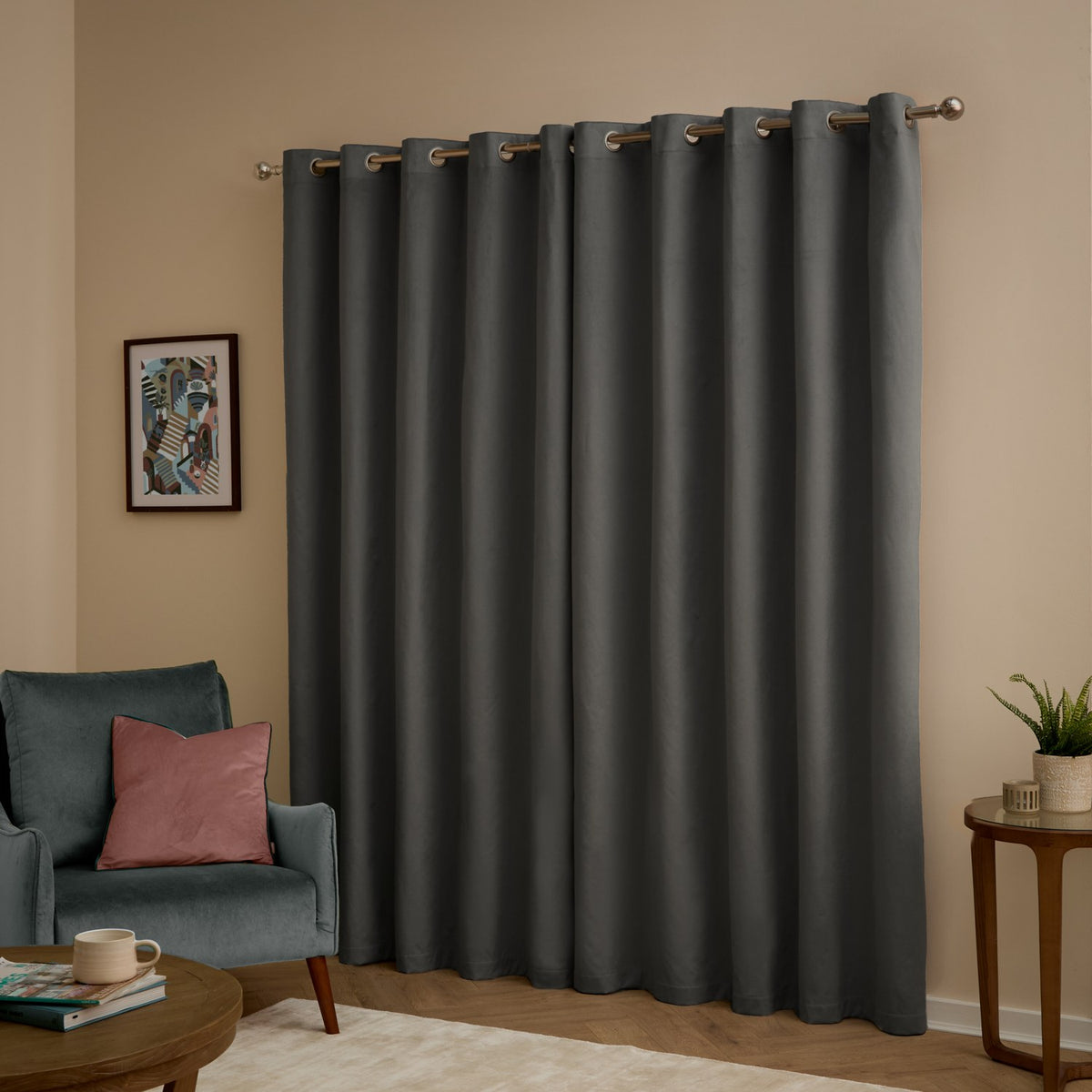 Charcoal grey velvet eyelet curtains with metal grommets hanging on a pole in a living room, shown with an armchair, side table, and framed wall art.