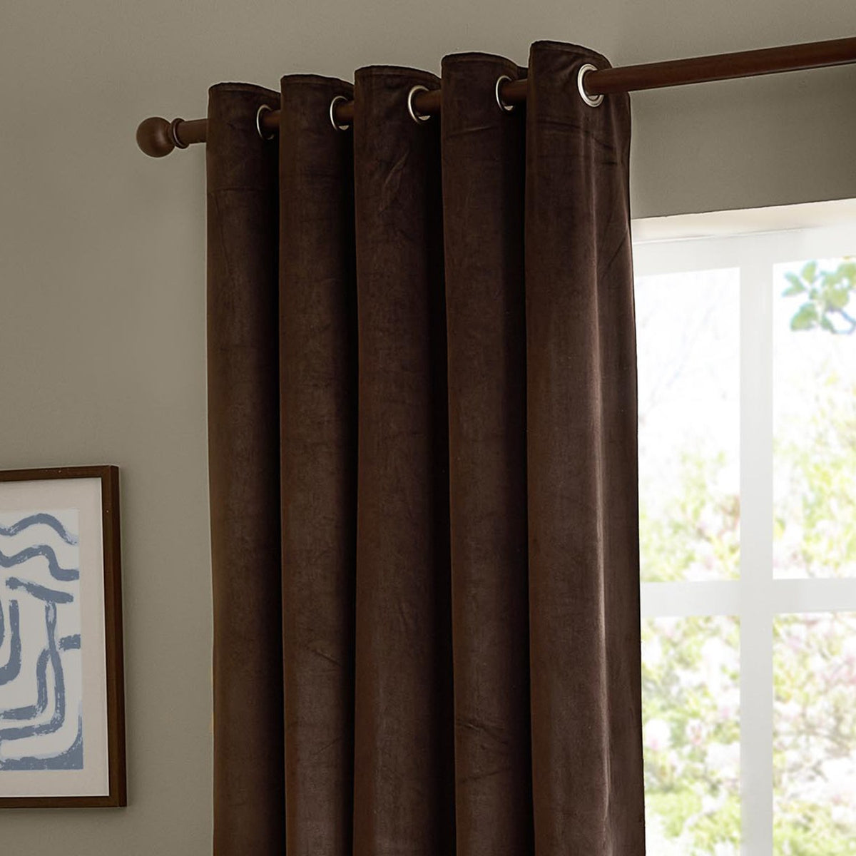 Chocolate-brown velvet eyelet curtains hanging on a wooden pole in a room, their metal grommets forming rounded folds beside a bright window.