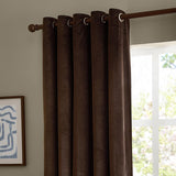 Chocolate-brown velvet eyelet curtains hanging on a wooden pole in a room, their metal grommets forming rounded folds beside a bright window.