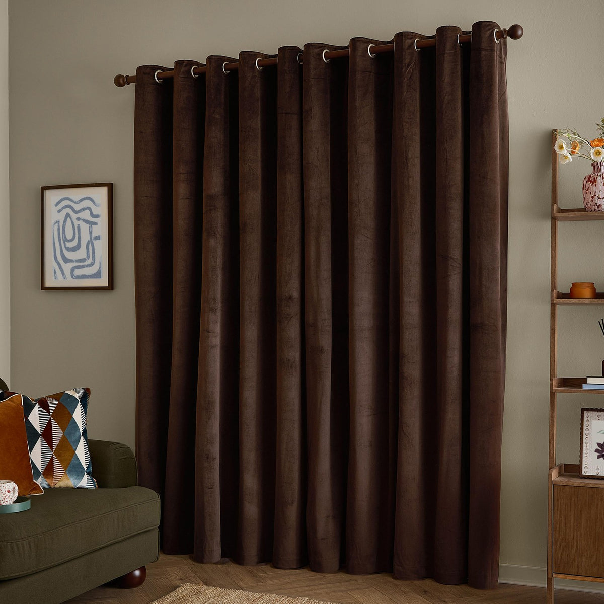Chocolate-brown velvet eyelet curtains hanging in soft folds on a wooden pole in a living room with a wall print and shelving unit.