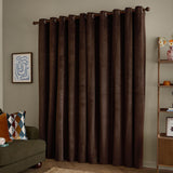 Chocolate-brown velvet eyelet curtains hanging in soft folds on a wooden pole in a living room with a wall print and shelving unit.