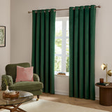 Emerald green velvet eyelet curtains hanging full-length either side of a window in a living room, styled with a green armchair and pink cushion.