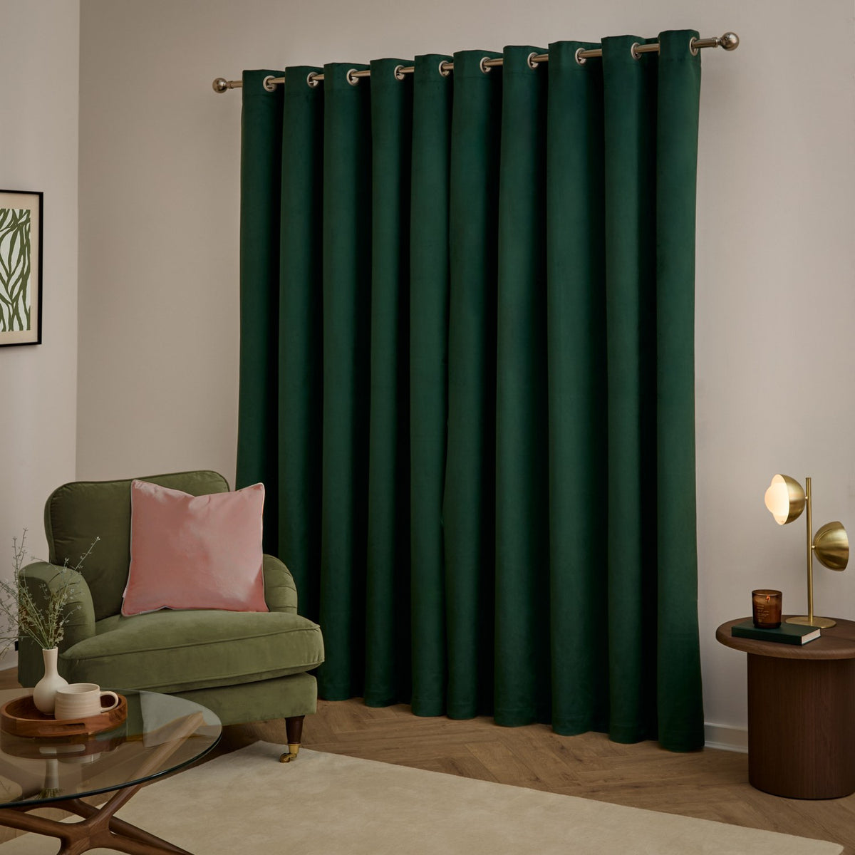 Emerald green velvet eyelet curtains with brushed metal rings, shown hanging on a pole in a living room with an olive armchair and pink cushion.