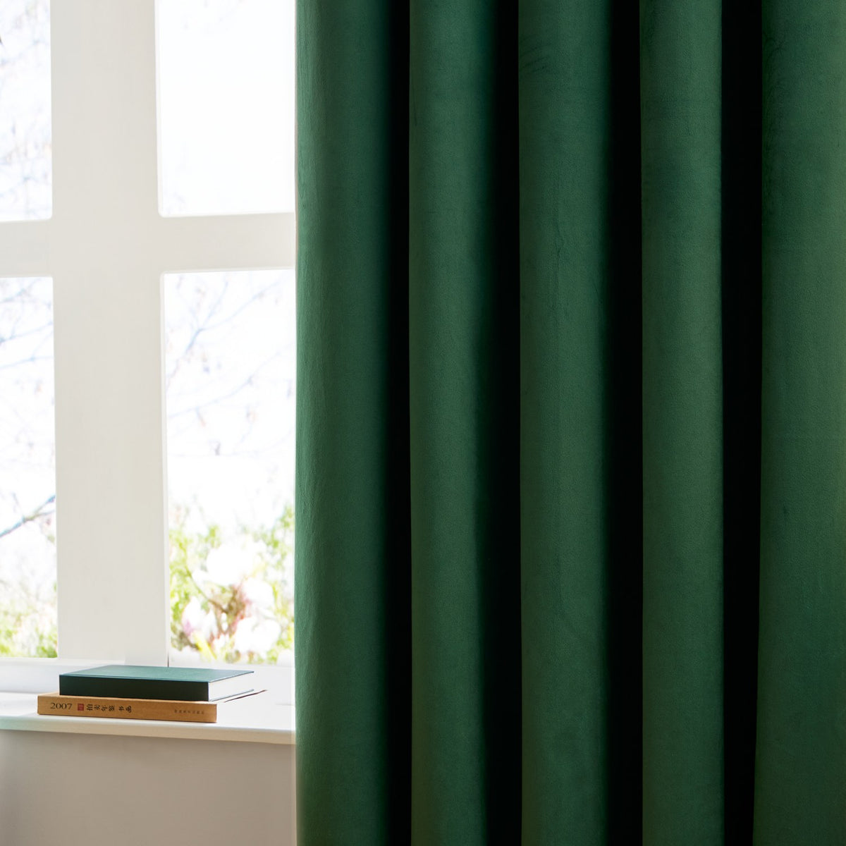 Emerald green velvet eyelet curtains hanging in folds beside a white-framed window, shown with a notebook on the sill in a bright living room setting.