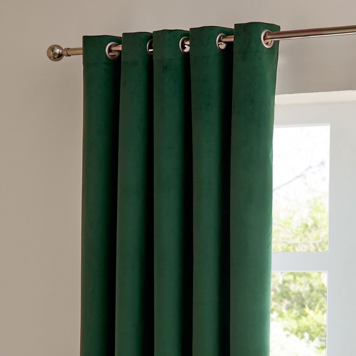 Emerald green velvet eyelet curtain in matt blackout fabric, shown hanging in soft folds from a chrome pole beside a window.