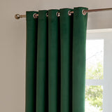 Emerald green velvet eyelet curtain in matt blackout fabric, shown hanging in soft folds from a chrome pole beside a window.