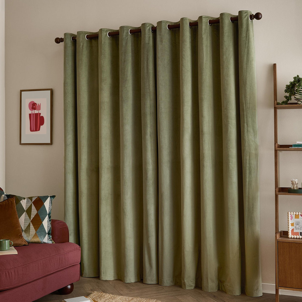 Sage green velvet eyelet curtains with grommet top hanging on a wooden rod, styled in a living room beside a red sofa and wooden shelving unit.