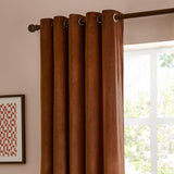 Rust-brown velvet eyelet curtains with metal grommets hung on a bronze pole against a pink wall beside a bright window.
