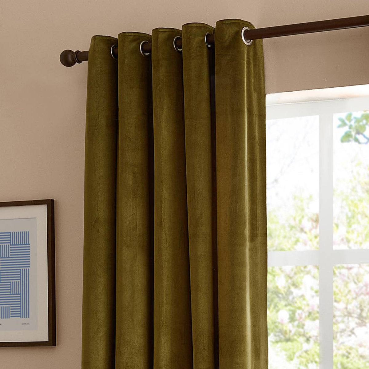 Olive velvet eyelet curtains with metal grommets on a dark pole, shown against a light pink wall beside a bright window.