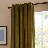 Olive velvet eyelet curtains with metal grommets on a dark pole, shown against a light pink wall beside a bright window.