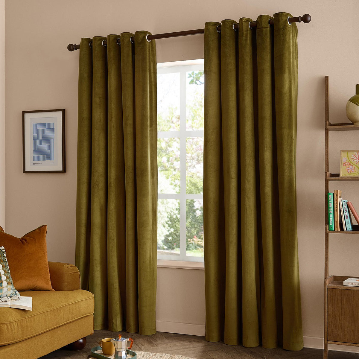 The Ultimate Velvet Plain Lined Eyelet Curtains in olive green hang from a dark eyelet pole in a living room, framing a window beside a mustard sofa and wooden shelving.