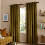 The Ultimate Velvet Plain Lined Eyelet Curtains in olive green hang from a dark eyelet pole in a living room, framing a window beside a mustard sofa and wooden shelving.