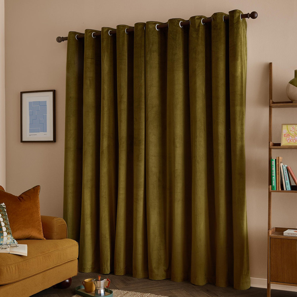Olive green velvet eyelet curtains with a matt finish hanging on a dark rod in a living room, styled with a mustard sofa and wooden shelving.