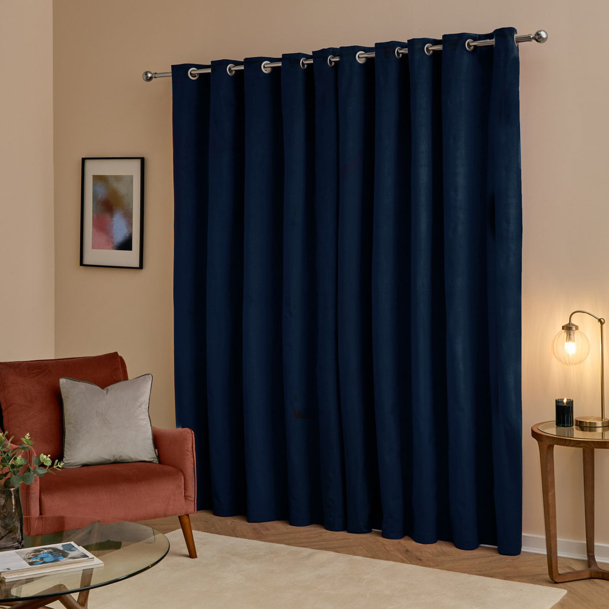 Navy blue velvet eyelet curtains with a fluid drape, hung on a silver pole in a living room with a rust armchair, glass table, and lit lamp.
