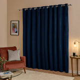 Navy blue velvet eyelet curtains with a fluid drape, hung on a silver pole in a living room with a rust armchair, glass table, and lit lamp.