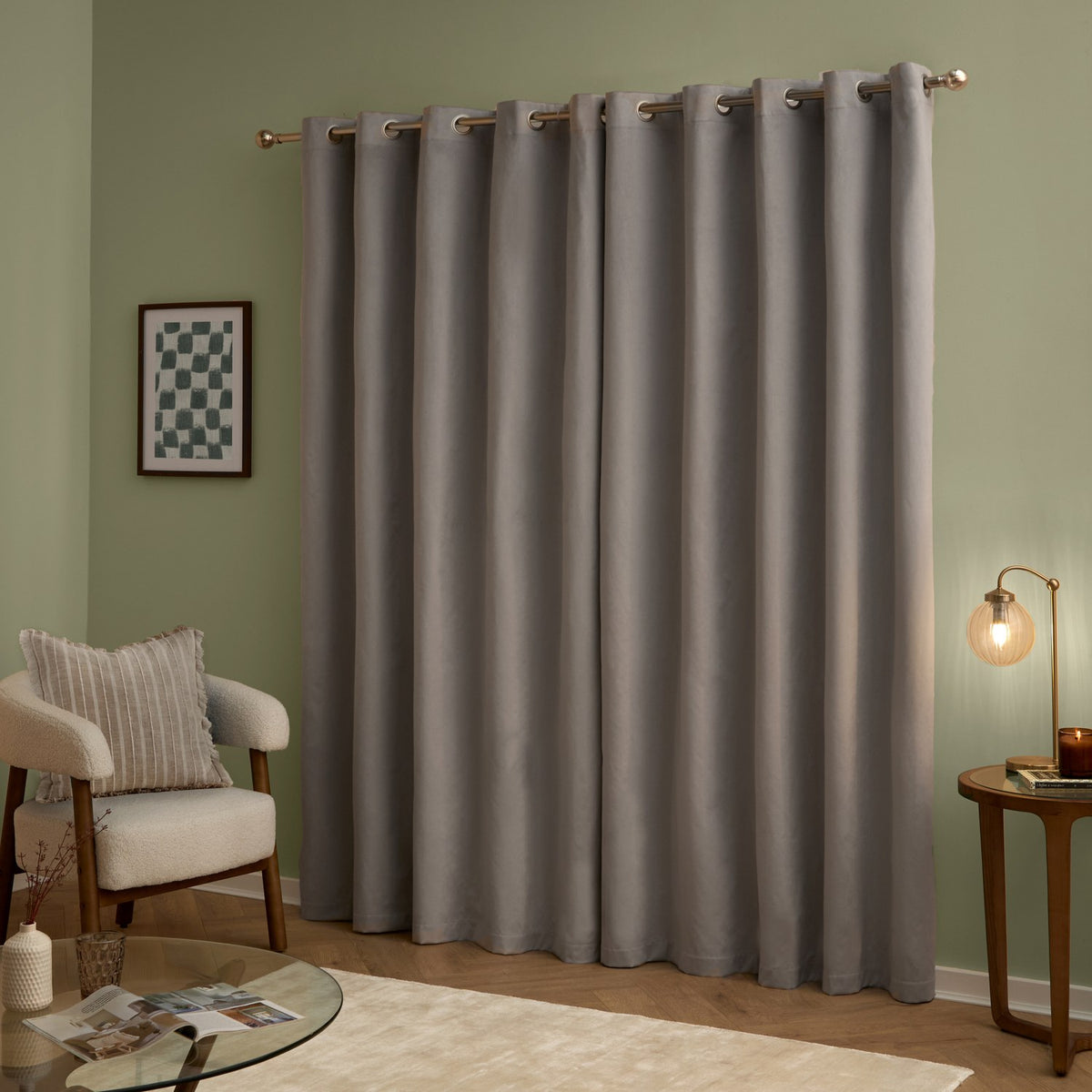 Silver velvet eyelet curtains hanging full-length from a metal pole in a green-walled living room, styled with an armchair, side table, lamp and glass coffee table.