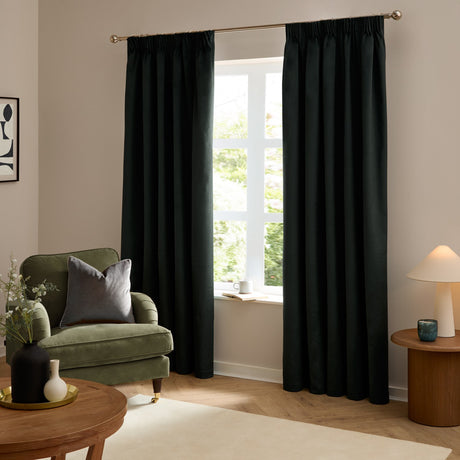 Black velvet pencil pleat curtains hanging full-length on a gold rod by a window, shown in a living room with a green armchair and wooden furniture.