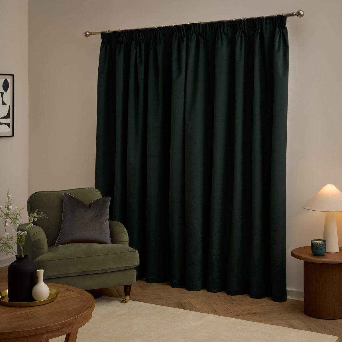 Dark green velvet pencil pleat curtains hanging full-length on a metal pole in a living room, shown with a green armchair, side table, and lamp.