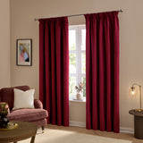 Burgundy velvet pencil pleat curtains hanging on a chrome pole either side of a window in a living room with a pink armchair and cushion.