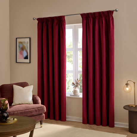 Burgundy velvet pencil pleat curtains hanging on a chrome pole either side of a window in a living room with a pink armchair and cushion.