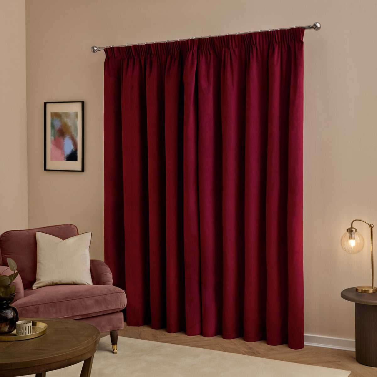 Burgundy velvet pencil pleat blackout curtains on a chrome pole, styled in a living room with a pink armchair, gold lamp and framed wall art.
