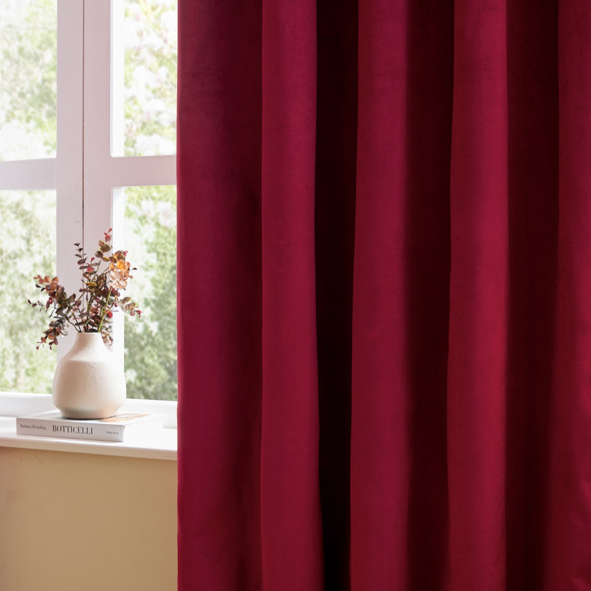 Burgundy velvet pencil pleat curtain with soft matt pile, shown pleated beside a window with a vase of dried flowers on the sill.