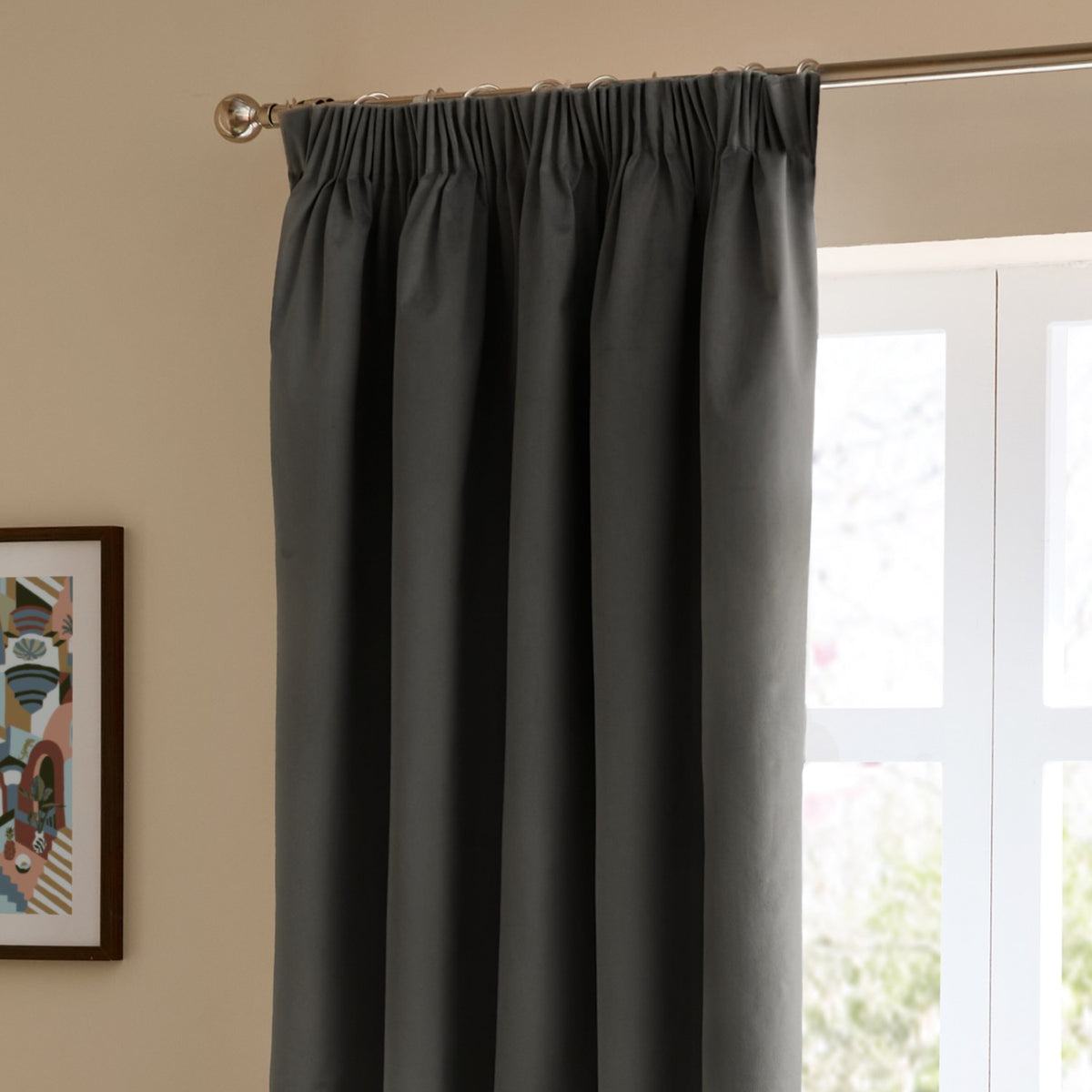 Charcoal grey velvet pencil pleat curtain hanging on a metal pole with clip rings, shown in a room beside a window and framed wall art.