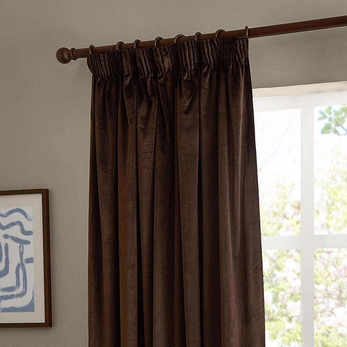 Chocolate brown velvet pencil pleat curtain with pencil pleat heading on a wooden pole, shown hanging beside a window in a living room setting.