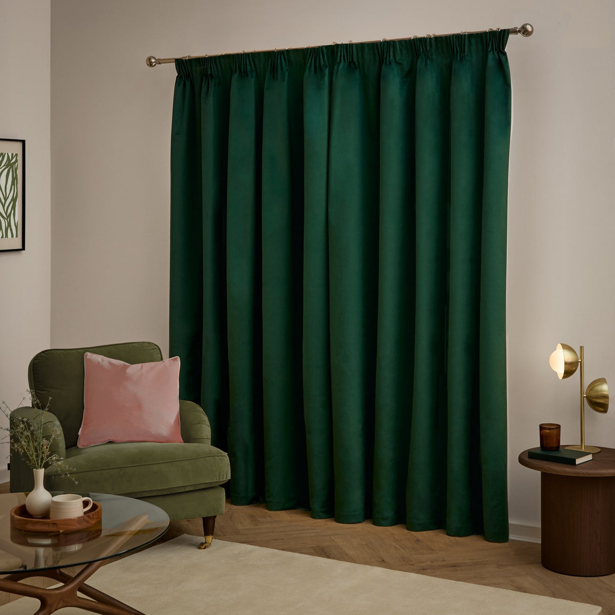 Emerald green velvet pencil pleat curtains with pencil pleat heading on a bronze pole, shown drawn in a living room with a green armchair, pink cushion, and wooden side table.