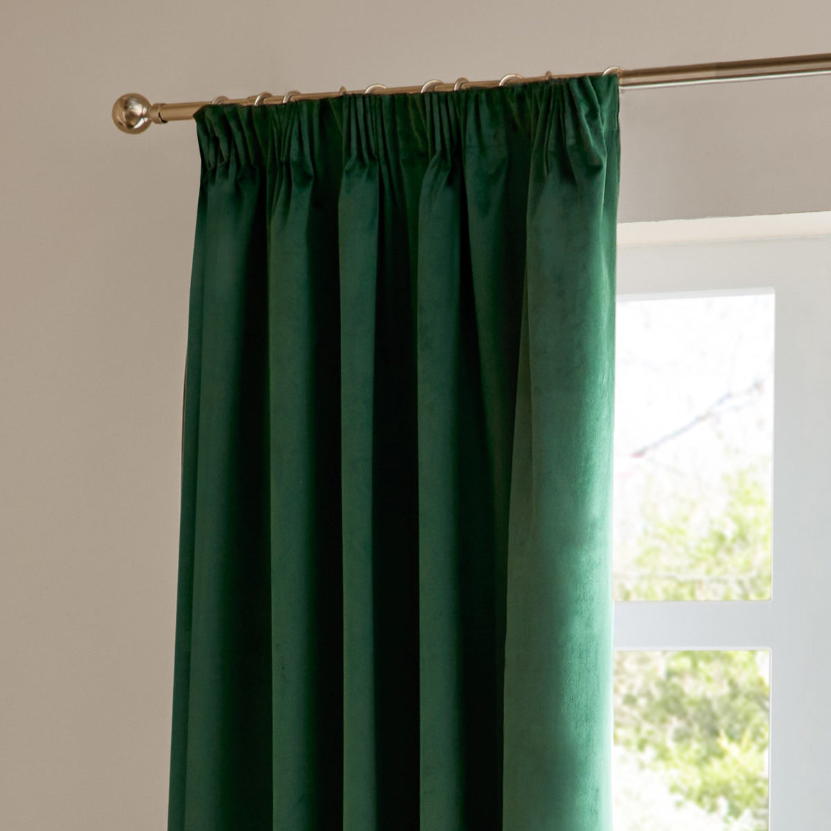 Emerald green velvet pencil pleat curtain hanging from a brass rod with pencil pleat tape and metal rings, shown beside a window.