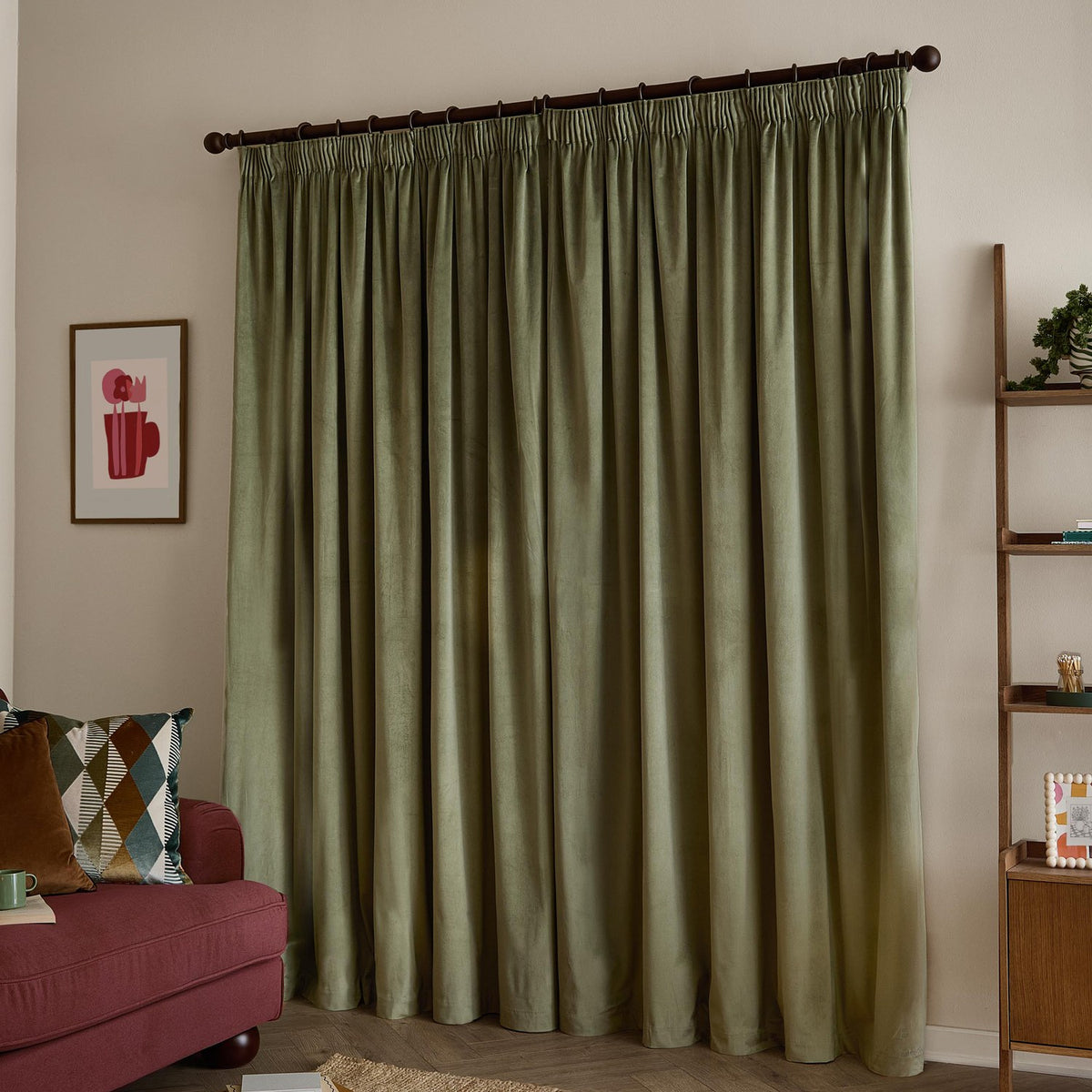 Sage green velvet pencil pleat curtains with pinch-pleated heading on a dark pole, shown pooling to the floor in a living room beside a red sofa.