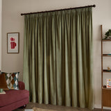 Sage green velvet pencil pleat curtains with pinch-pleated heading on a dark pole, shown pooling to the floor in a living room beside a red sofa.