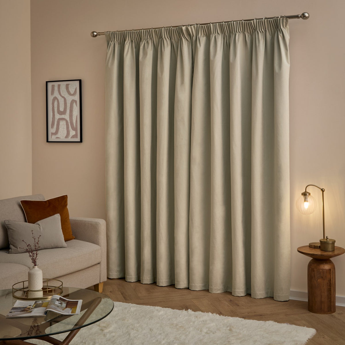 Natural velvet pencil pleat blackout curtains hung on a chrome rod in a living room with pink walls, wood flooring, and a beige sofa.