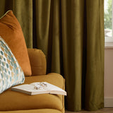 Olive green velvet pencil pleat curtains in soft folds beside a mustard sofa with an orange cushion, patterned pillow, and a book with glasses.