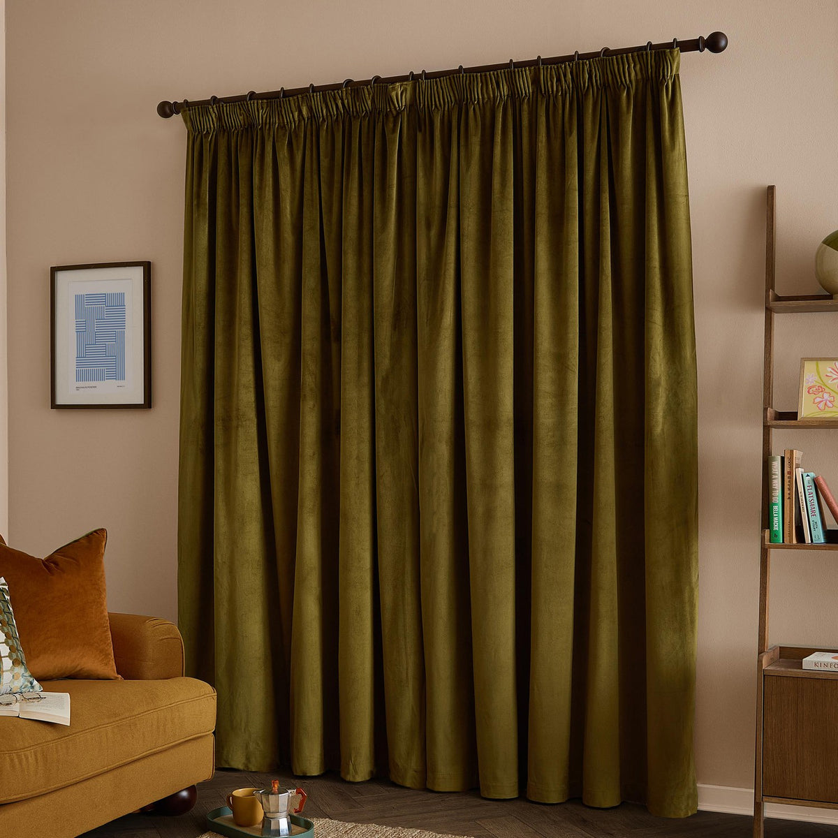 Olive green velvet pencil pleat curtains with pencil pleat heading on a dark rod, shown drawn across a window in a living room with a mustard sofa and shelving.