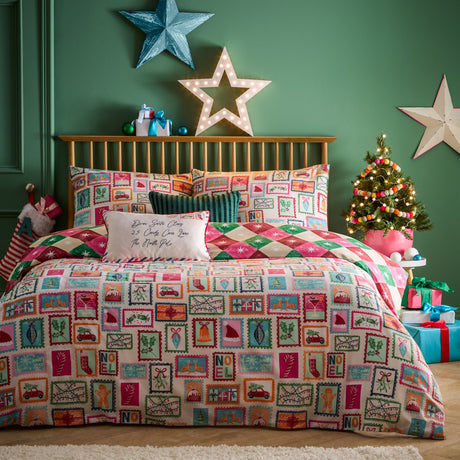 Dear Santa reversible duvet cover set on a bed, front side showing multicolour Christmas postage-stamp print with holly, cars, baubles and NOEL text on cream.