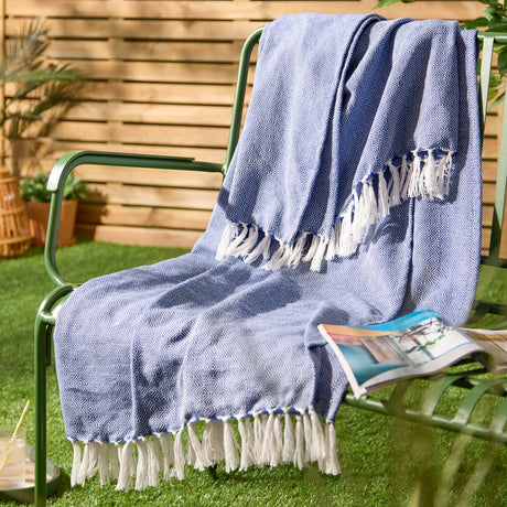 Dessa Geometric Polyester Throw in cobalt blue with white knotted fringing, draped over a green outdoor chair with a wooden slatted fence backdrop.