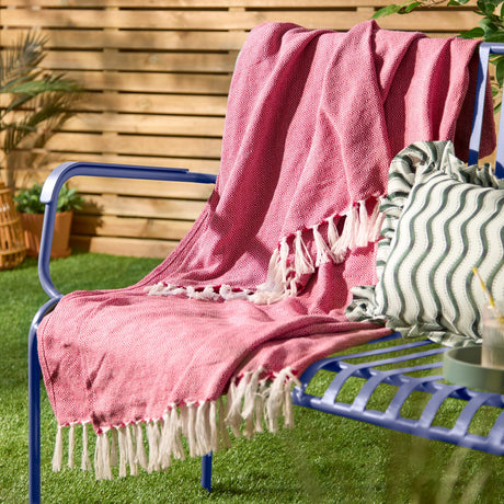 Dessa Geometric Polyester Throw in fuchsia draped over a blue outdoor bench, showing the diamond weave pattern and off-white knotted fringe.