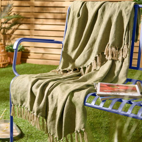 Dessa Geometric Polyester Throw in olive draped over a blue outdoor bench, showing diamond lattice pattern and knotted tassel fringe on a garden lawn setting.