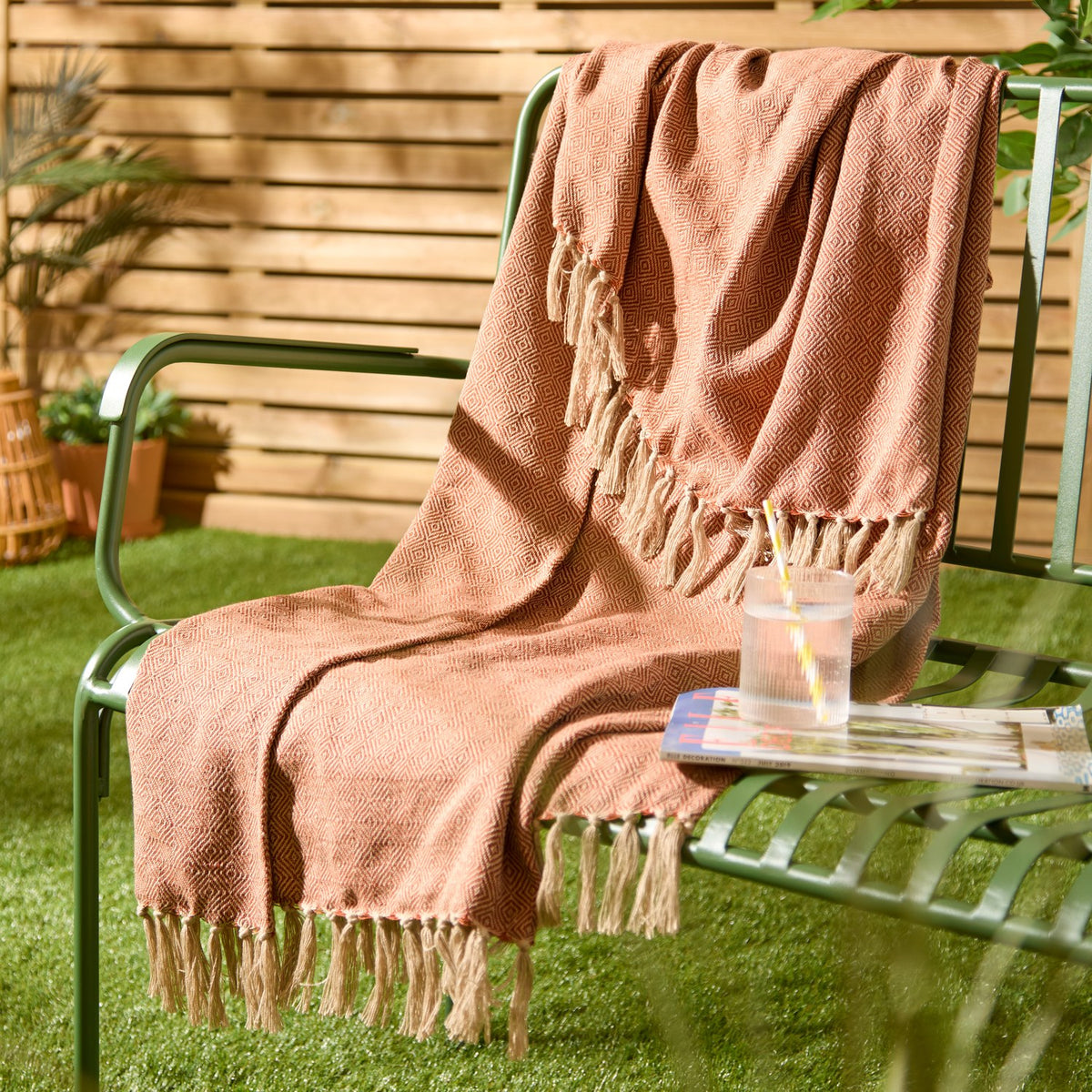 Dessa Geometric Polyester Throw in terracotta with cream diamond pattern and knotted fringe, draped over a green garden bench beside a drink and magazine.
