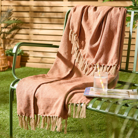 Dessa Geometric Polyester Throw in terracotta with cream diamond pattern and knotted fringe, draped over a green garden bench beside a drink and magazine.