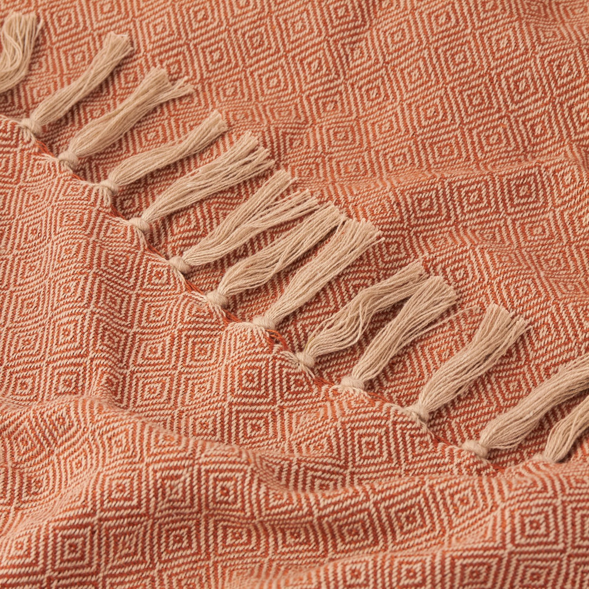 Close-up of the Dessa Geometric Polyester Throw in terracotta, showing a small diamond weave pattern with a knotted cream fringe along one edge.