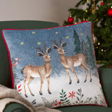 Ditsy Festive Reindeer Animal Velvet Cushion in multicolour, printed with two reindeer in a snowy scene and red piping, shown on a green armchair beside a Christmas tree.