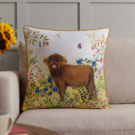 Daisy Hill Agnes velvet cotton cushion in multi/tan, printed with a Highland cow amid meadow flowers, cornflowers and a butterfly, shown on a beige sofa.