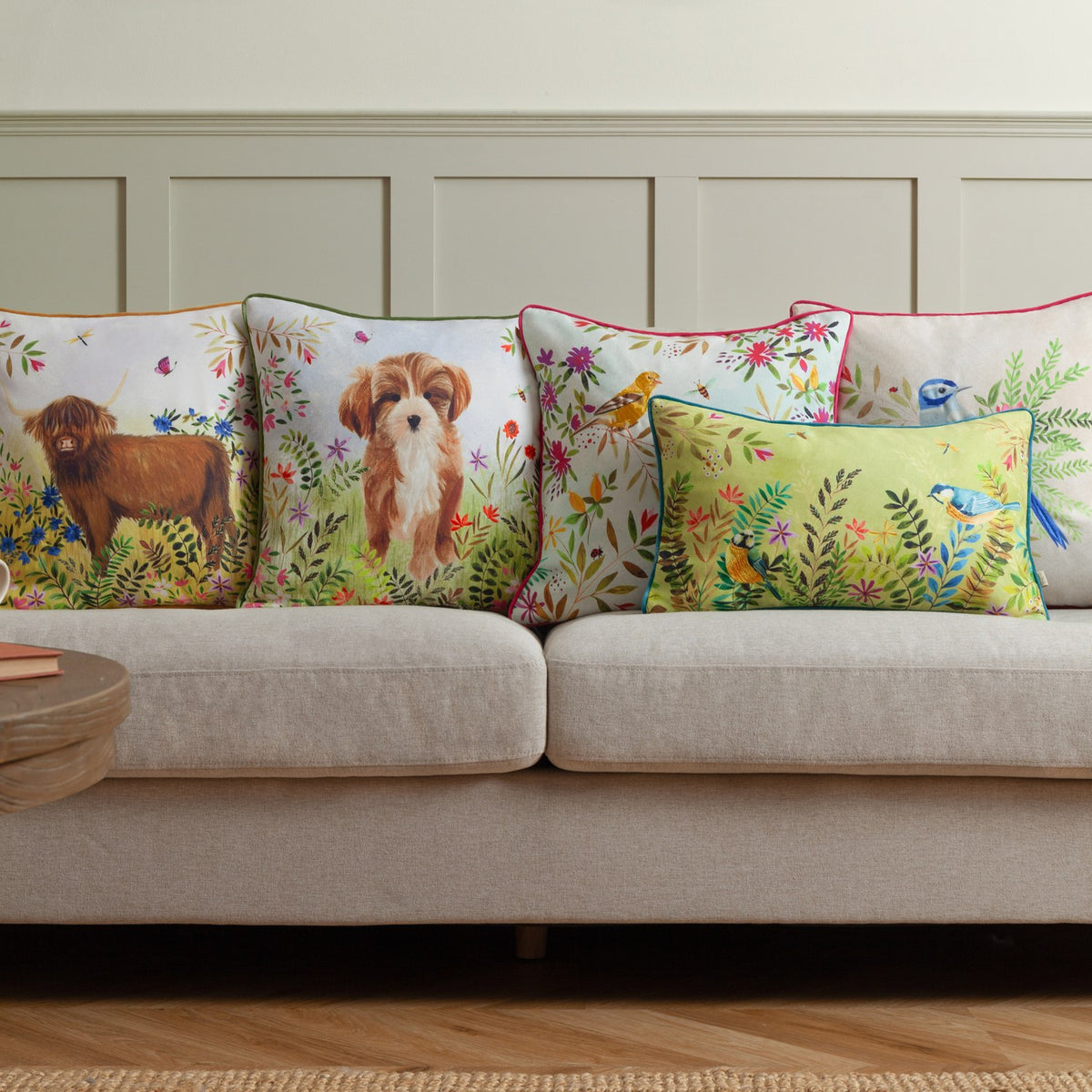 Square scatter cushion in the Daisy Hill Agnes design, printed with a Highland cow in a meadow of leaves and flowers, shown on a beige sofa among coordinating animal and bird cushions.
