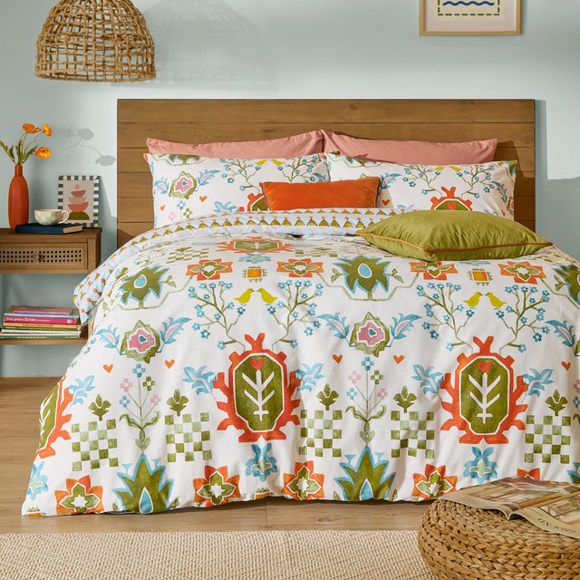 Dilara Reversible Geometric Duvet Cover Set styled on a bed with folk-art bird and floral print in orange, olive, blue and pink on cream cotton-blend fabric, shown in a light blue bedroom.