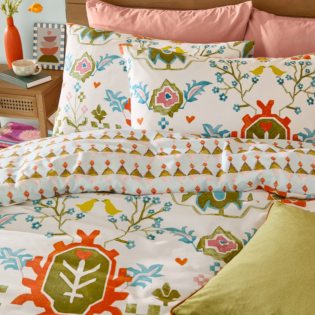 Dilara Reversible Geometric Duvet Cover Set on a bed, featuring folk-art birds and floral motifs in orange, olive, blue and pink on cream fabric, styled with pink and green cushions.